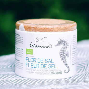 Jar of Flor de Sal with belamandil branding on a textured surface with green blurred background
